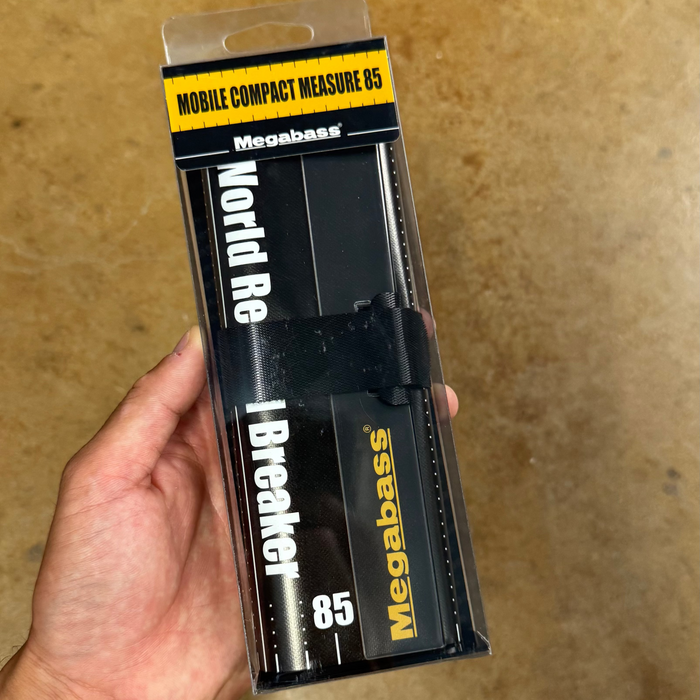 Hand holding a Megabass branded fishing reel package on a brown surface